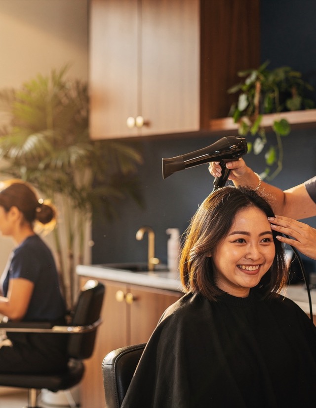 A stylist blow-drying a smiling client’s hair, warm salon light